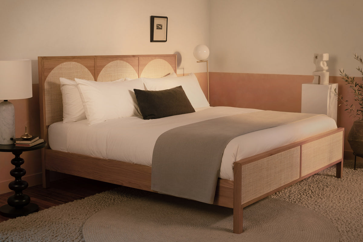 A bedroom with a contemporary wood and rattan bed made with LETTO Americano Cotton Percale sheets, a gray blanket folded at the foot of the bed, and a charcoal velvet throw pillow. data-image-id=