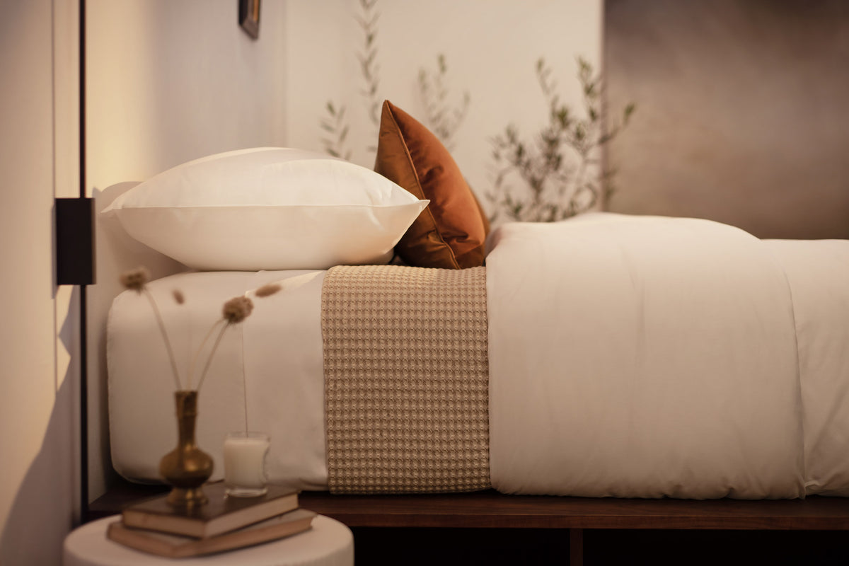 A close up picture of a bed made with LETTO White Classic Cotton Sateen sheets and duvet cover, with a Nido waffleweave blanket in beige and an orange velvet throw pillow.  data-image-id=