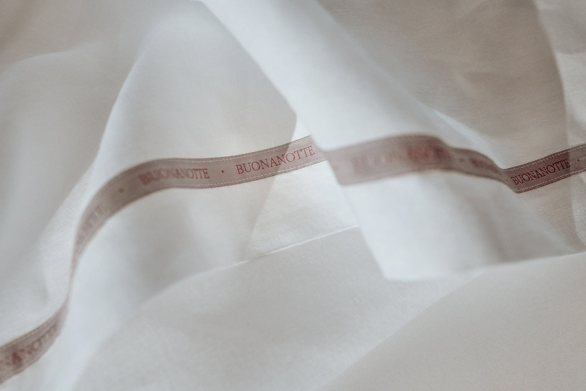 A close up image of the Buonanotte ribbon on the bottom edge of a breathable and relaxed 100% linen LETTO Lino Primo flat sheet in color white. data-image-id=