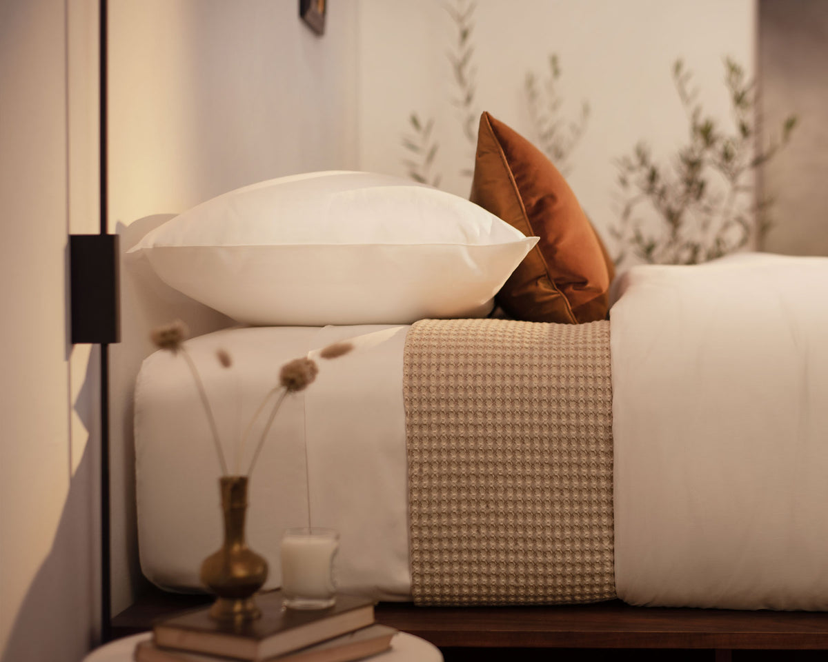 A close up picture of a bed made with LETTO White Classic Cotton Sateen sheets and duvet cover, with a Nido 100% Cotton waffleweave blanket in beige and an orange velvet throw pillow. data-image-id=