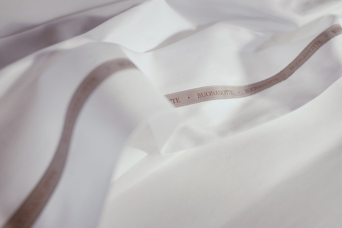 A close up image of the Buonanotte ribbon on the bottom edge of buttery and transcendent LETTO Sea Island Cotton Sateen flat sheet in color white. data-image-id=
