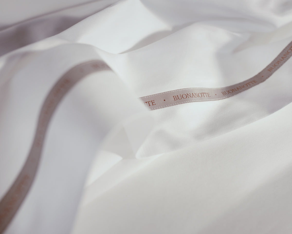 A close up image of the Buonanotte ribbon on the bottom edge of buttery and transcendent LETTO Sea Island Cotton Sateen flat sheet in color white. data-image-id=