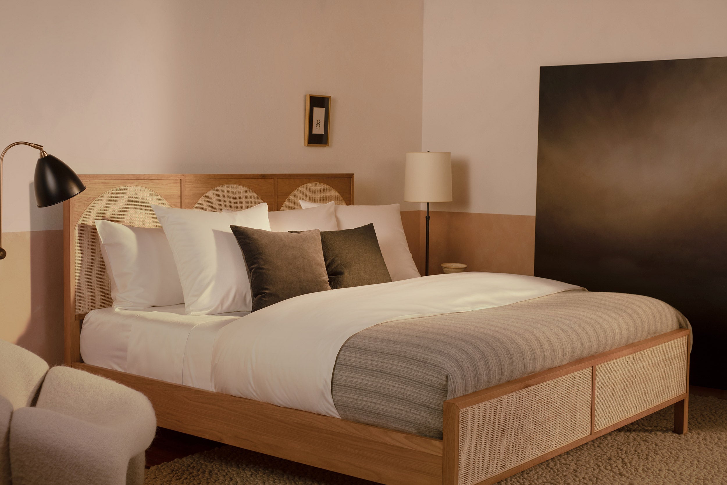A bedroom with a contemporary wood and rattan bed made with LETTO Americano Cotton Sateen sheets, a striped gray duvet cover, and charcoal velvet throw pillows.