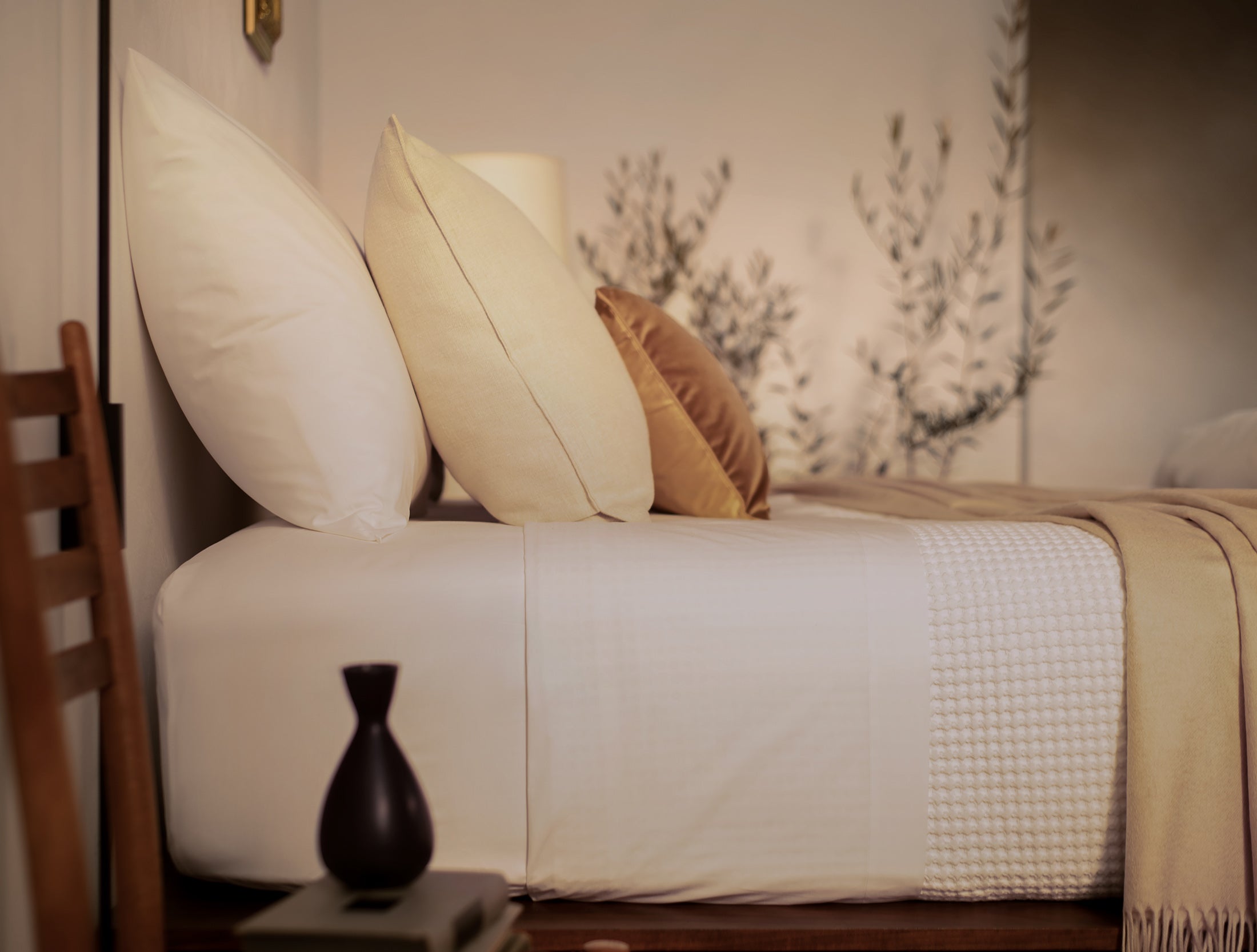 A close up picture of a bed made with LETTO White Classic Cotton Percale sheets and duvet cover, with a Nido waffleweave blanket in white and a tan velvet throw pillow.