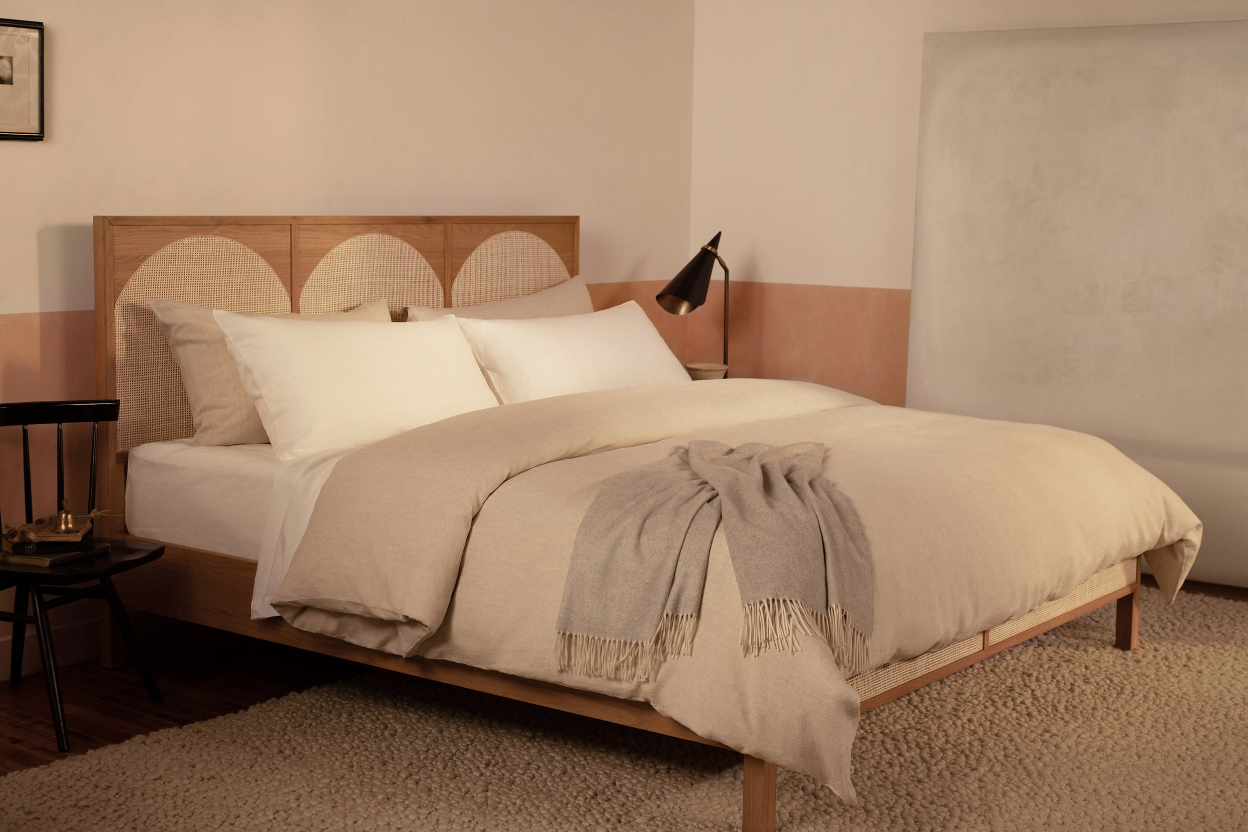 A bedroom with a contemporary wood and rattan bed made with LETTO Classic Linen sheets in white and Classic Linen Duvey in natural, with a gray Coccola cashmere throw blanket.