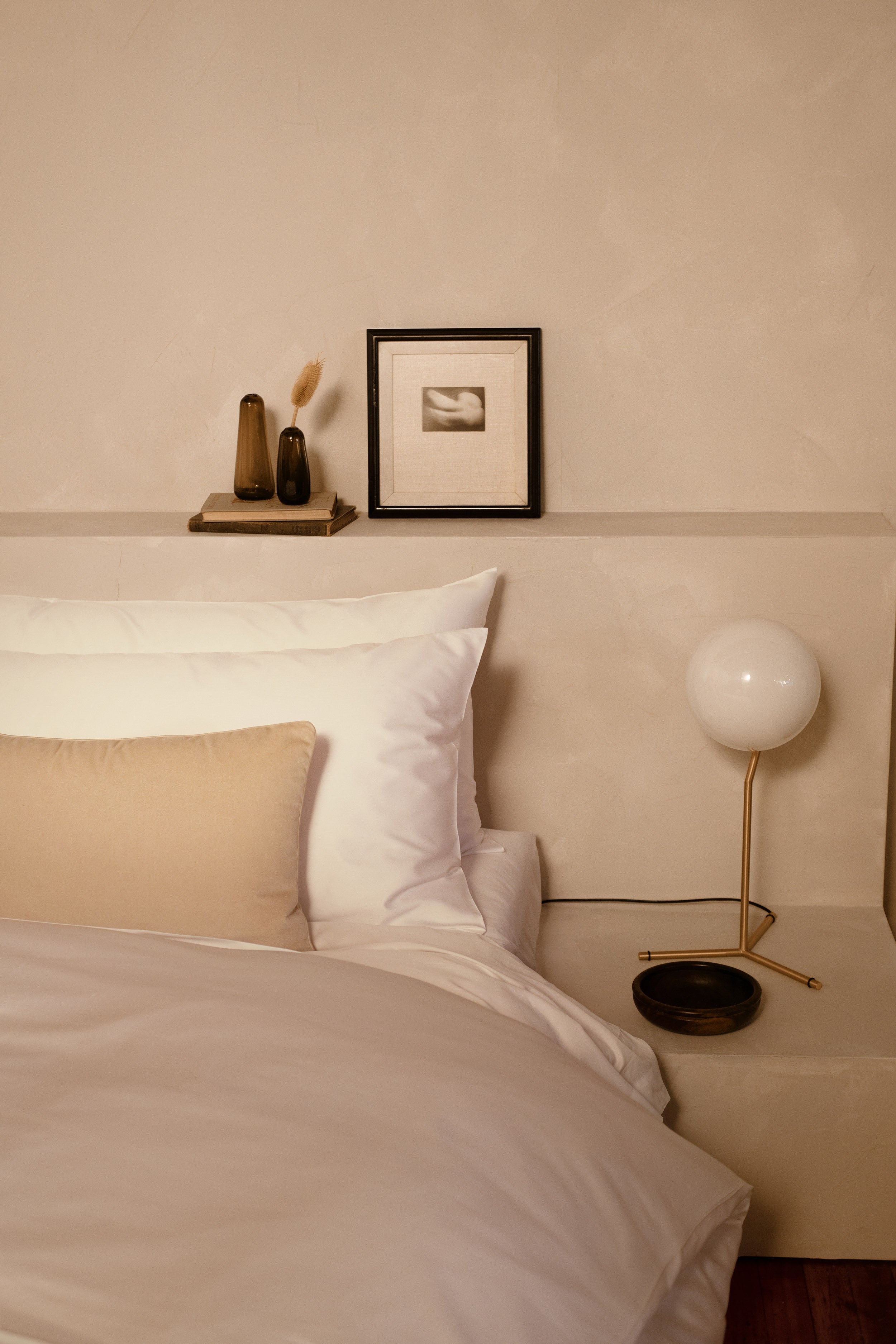 A close up image of LETTO Giza Reserve Cotton pillows in white on a bed with a tan velvet throw pillow and a nightstand with a lamp.
