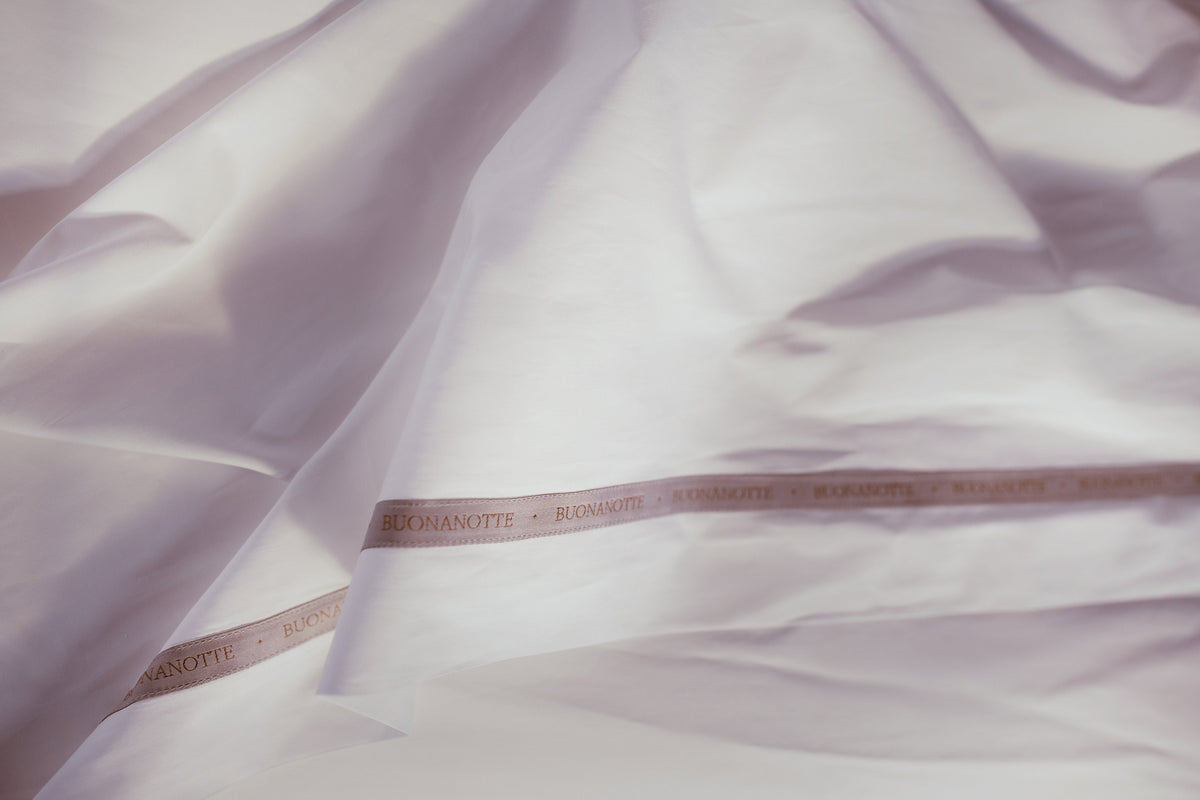 A close up image of the Buonanotte ribbon on the bottom edge of crisp and cool LETTO Americano Cotton Percale flat sheet in color white. data-image-id=