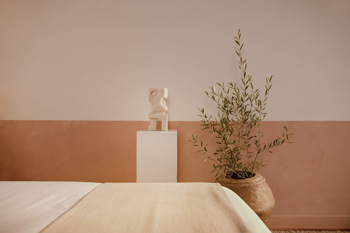 An image of the foot of a bed with an small olive tree in a planter againsts a rustic plaster wall next to a small statue. data-image-id=