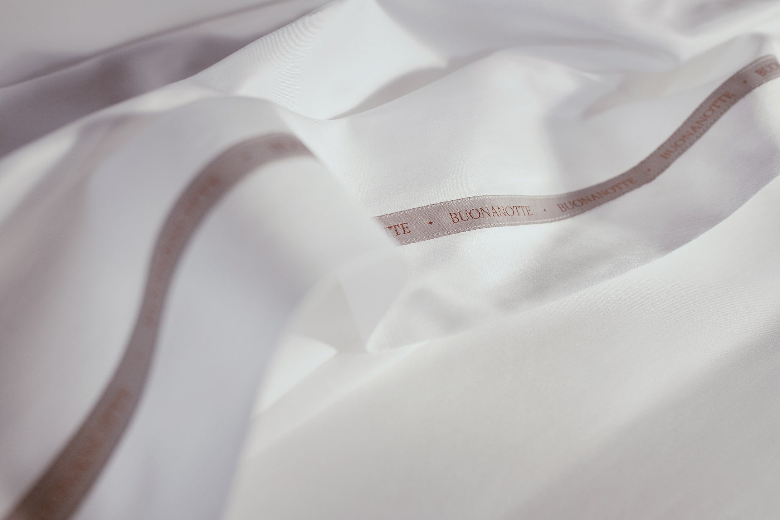 A close up image of the Buonanotte ribbon on the bottom edge of smooth and buttery LETTO Americano Cotton Sateen flat sheet in color white.