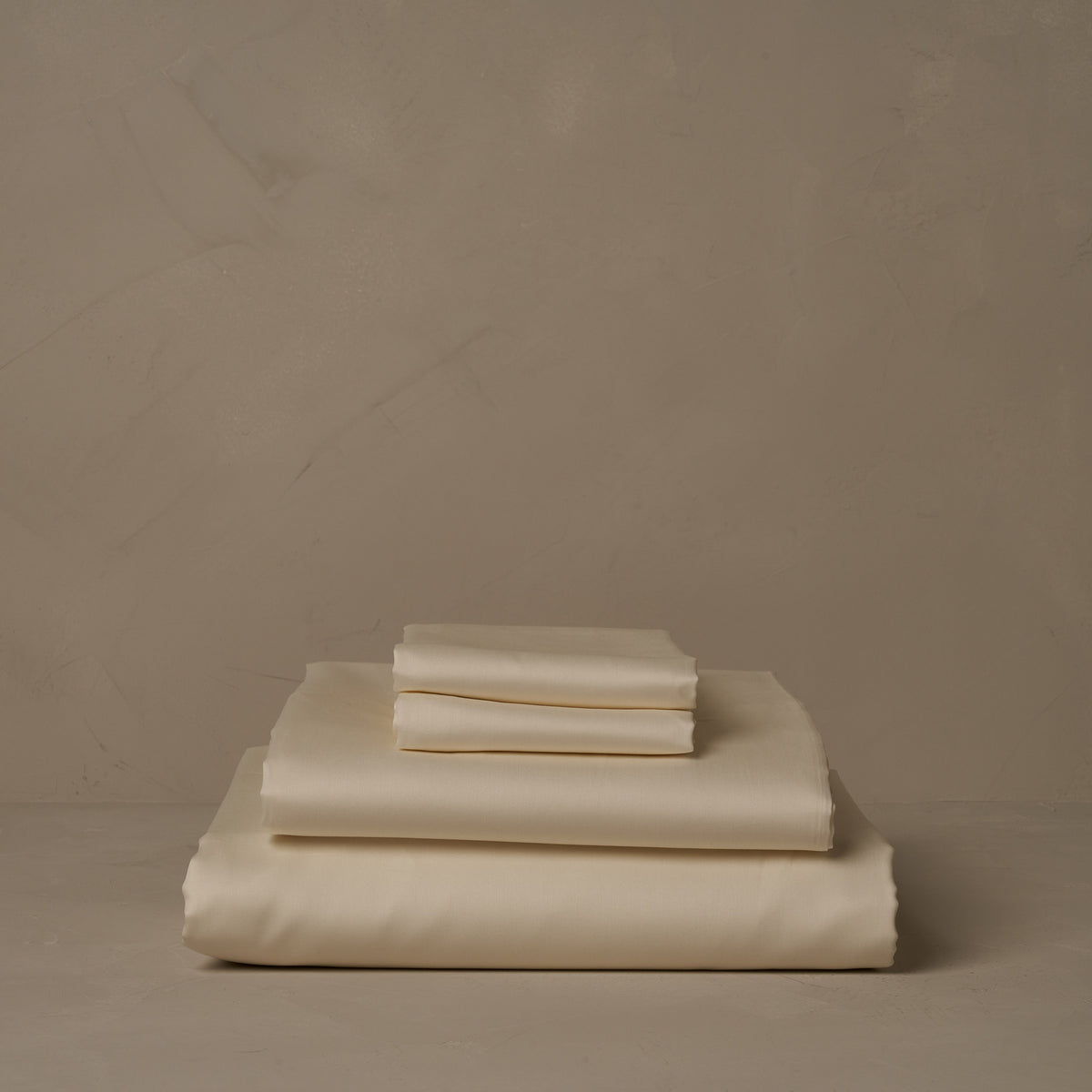 A stack of warm and buttery Classic Cotton Sateen sheet sets in ivory, made in Italy. The sheet set includes a fitted sheet, a flat sheet, and a pair of pillowcases. data-image-id=