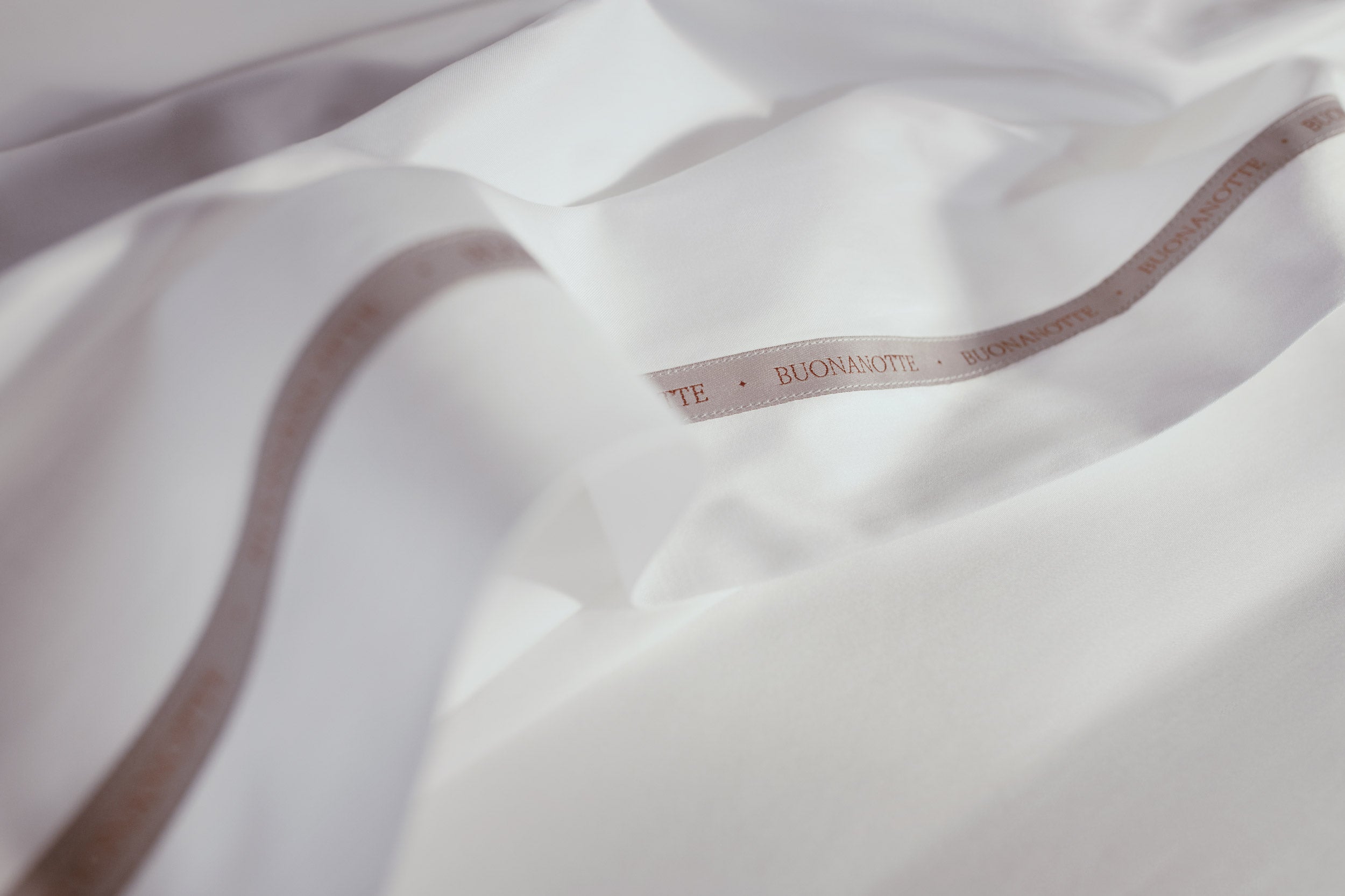 A close up image of the Buonanotte ribbon on the bottom edge of buttery and transcendent LETTO Sea Island Cotton Sateen flat sheet in color white.