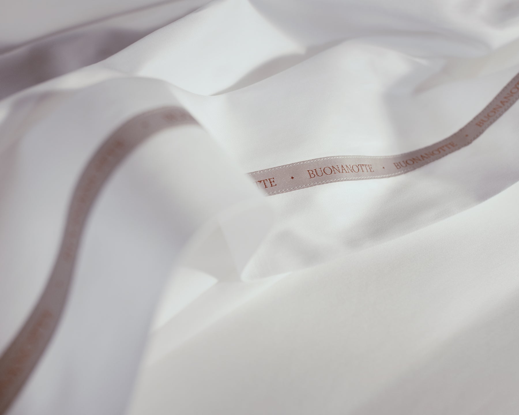 A close up image of the Buonanotte ribbon on the bottom edge of buttery and transcendent LETTO Sea Island Cotton Sateen flat sheet in color white.